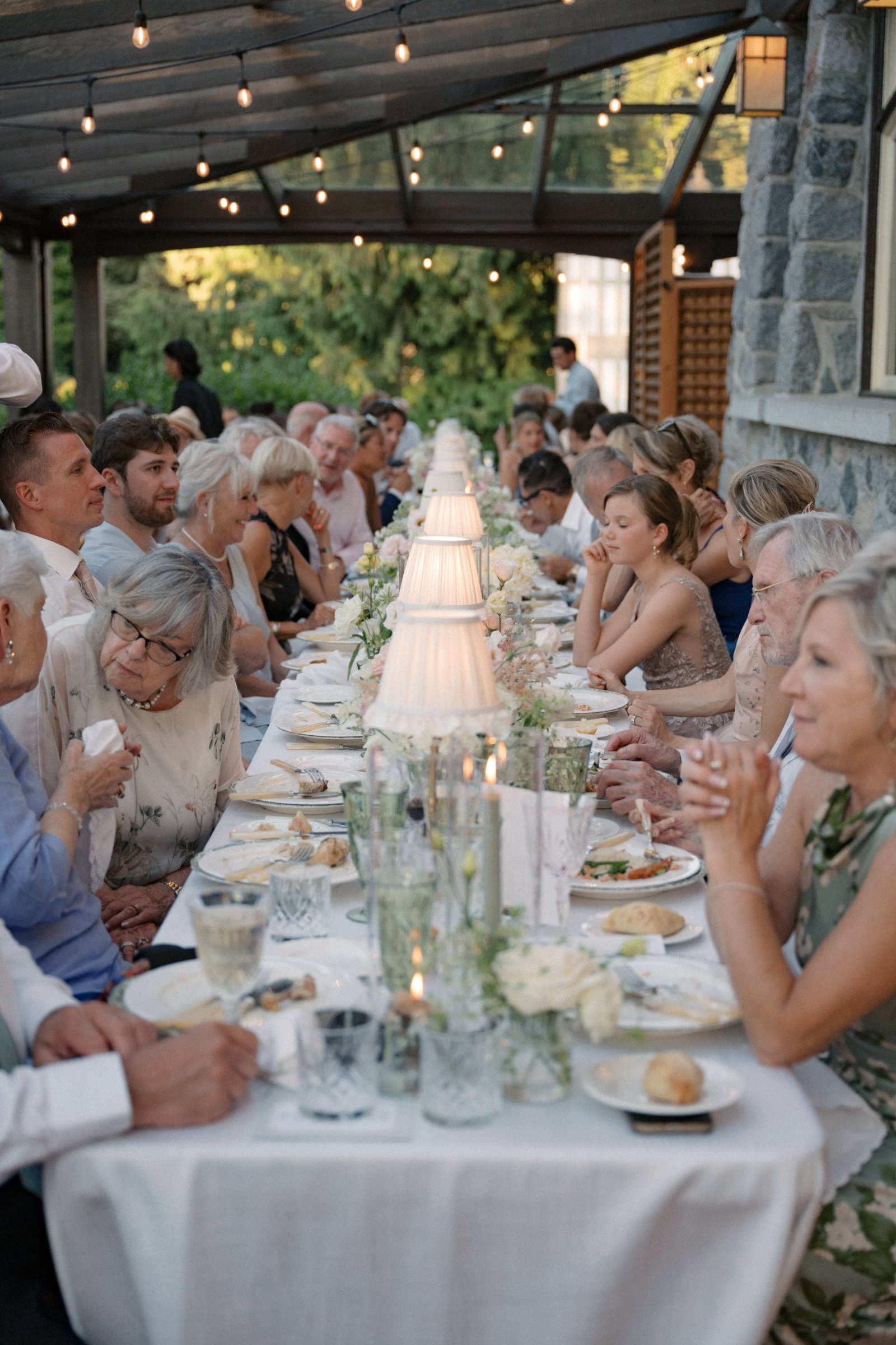 Guests dining beneath brass table lamps with crème ruffled shades along an elegant outdoor reception table with lush florals and candlelight.