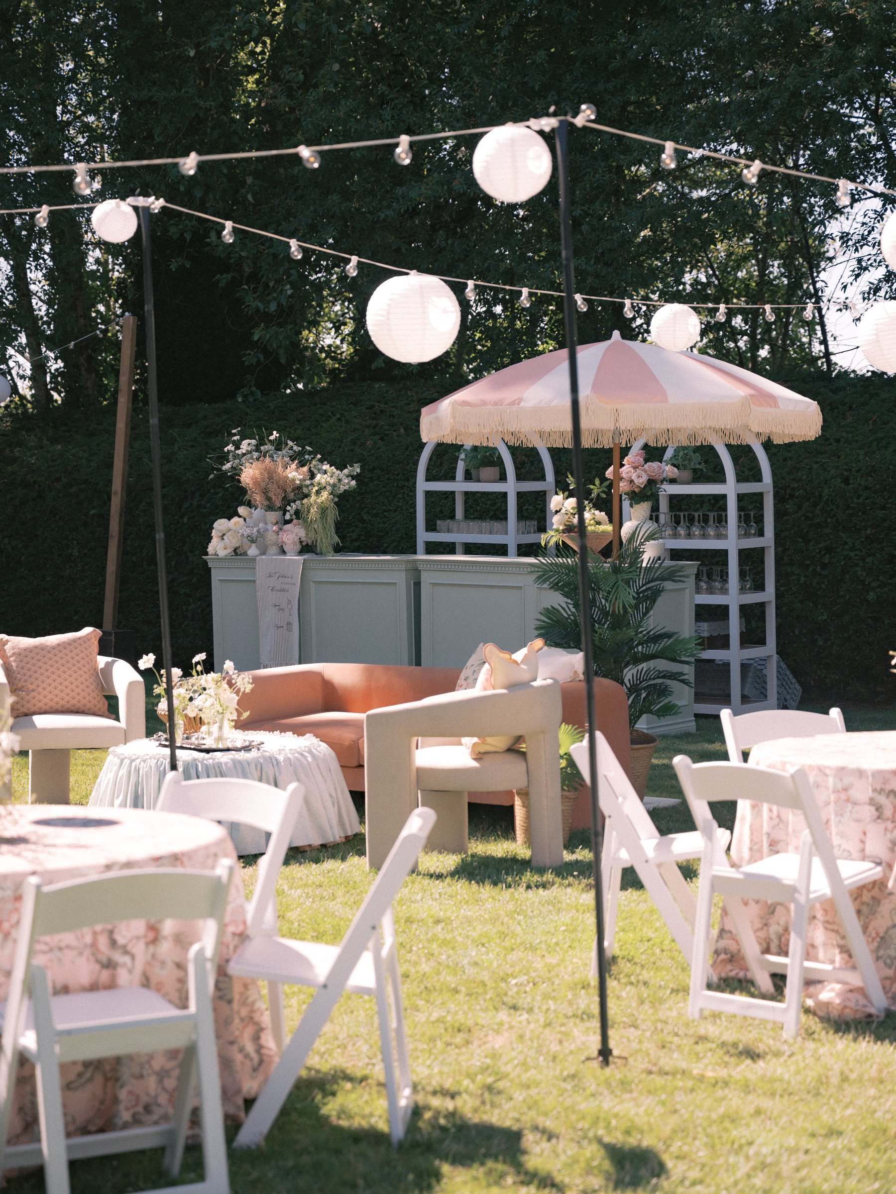 Elegant outdoor wedding lounge featuring a pastel pink and white market umbrella, blush seating, café tables, and romantic garden party styling.