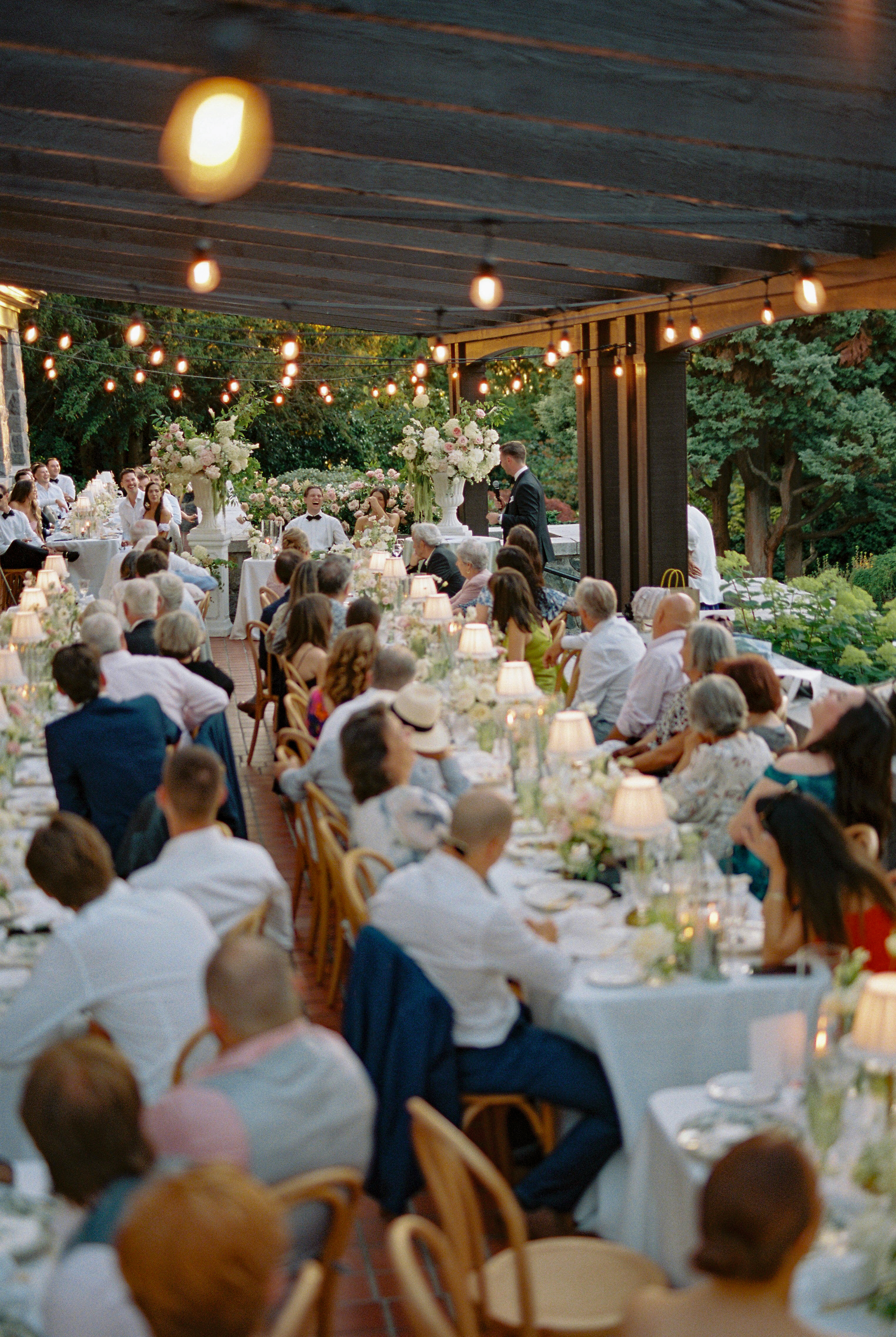 Outdoor evening wedding reception illuminated by brass table lamps with crème ruffled shades, candlelight, and romantic string lights.